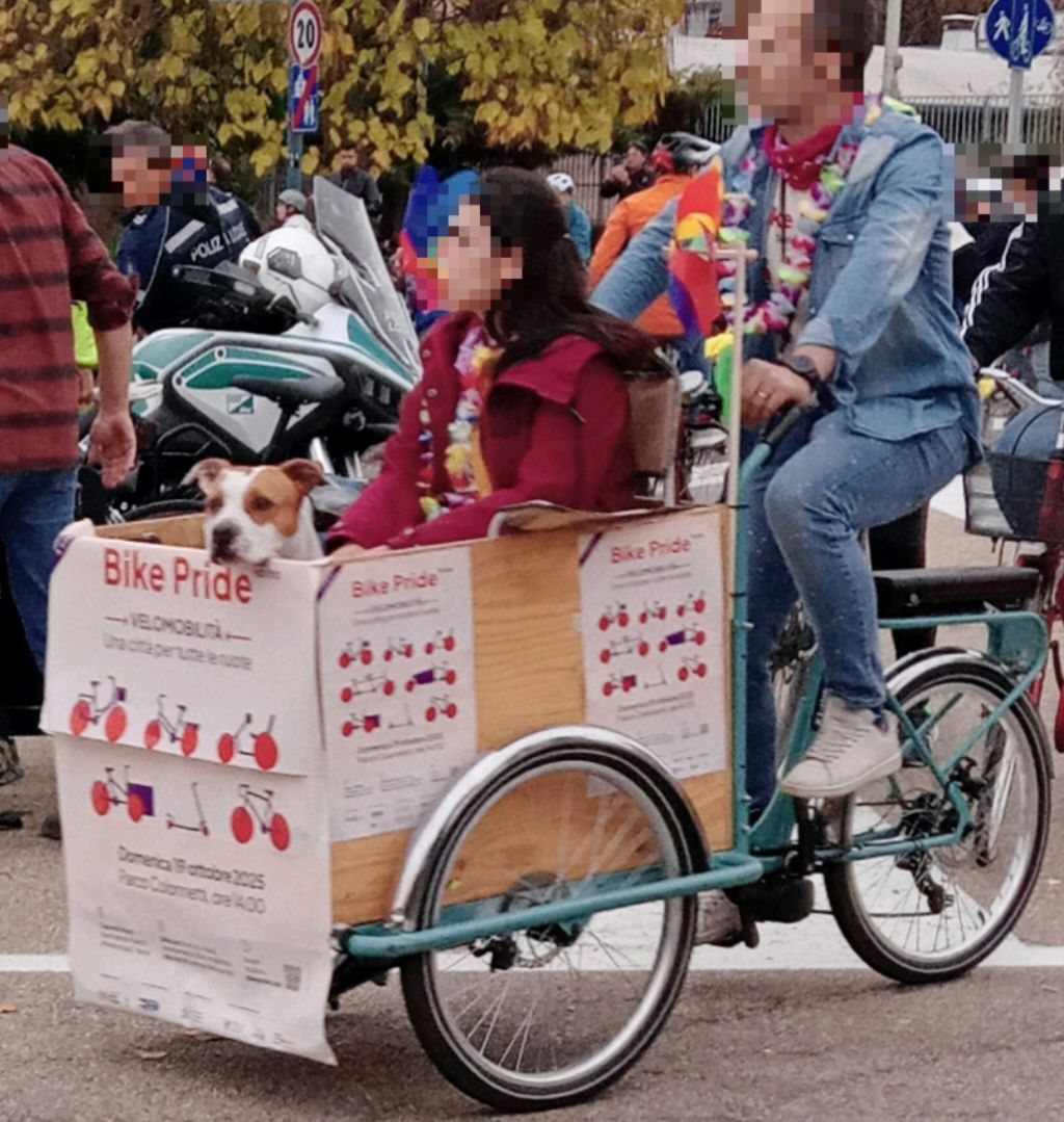 Nel cassone di una bici cargo, sta seduta una ragazza accanto alla quale si affaccia un cane. Il cassone è tappezzato di manifesti dell'iniziativa "Bike Pride" del 2025.